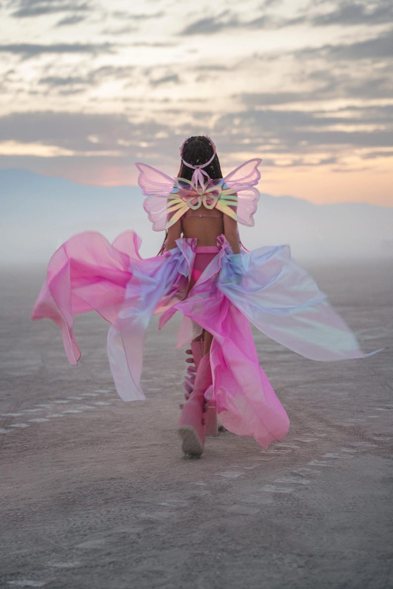 Puede incluir: Una persona vestida con un disfraz de mariposa rosa e iridiscente con alas y una falda fluida, caminando sobre una superficie blanca y polvorienta.