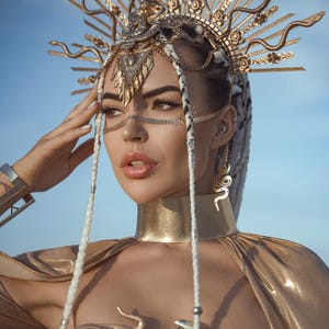 Puede incluir: Tocado dorado con diseños de serpientes y rayos de sol. La modelo lleva un collar gargantilla dorado, pendientes a juego y un top dorado. Trenzas blancas adornadas con cuentas plateadas. El fondo es un cielo azul claro.