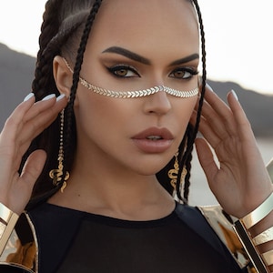 Puede incluir: Una mujer con el pelo negro trenzado lleva una cadena dorada sobre la frente y pendientes de serpiente a juego. Lleva una camiseta de malla negra con detalles dorados y un brazalete dorado.