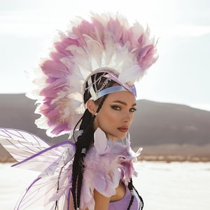 以下が含まれることがあります： ピンクと白の羽飾り、ラベンダー色のボディスーツ、妖精の羽が特徴のファンタジーコスチューム。衣装には銀色の装飾が施され、砂漠の風景を背景にしています。テーマのあるイベントに最適です。