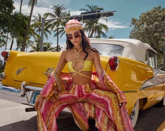 Puede incluir: Una mujer con un top amarillo de halter con un detalle de anillo dorado, pantalones a rayas rosa y amarillo y un pañuelo estampado en rosa y amarillo. Está sentada en la parte trasera de un coche amarillo con molduras blancas.
