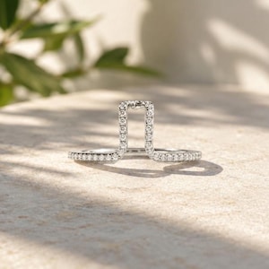 Peut inclure: Une bague au design moderne et ouvert, avec une partie centrale rectangulaire et deux anneaux, tous incrustés de petites pierres scintillantes. La bague est posée sur une surface claire, avec des ombres douces et un arrière-plan vert flou.