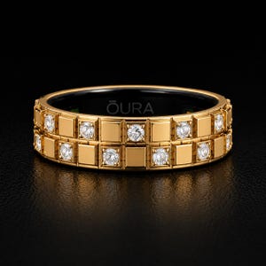 Peut inclure: Une bague dorée présentant un motif géométrique à double rangée composé de plaques métalliques carrées alternées avec des pierres claires rondes. L'intérieur de l'anneau est noir et affiche le texte OURA en blanc, centré dans la structure.