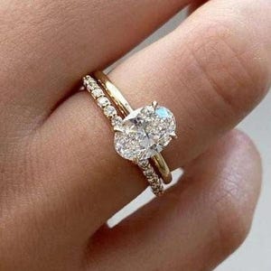 Puede incluir: Una banda de oro con una fila de pequeños diamantes se encuentra debajo de un anillo de diamantes de forma ovalada más grande con una banda de oro.