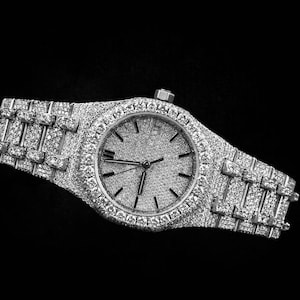 Könnte beinhalten: Eine luxuriöse Armbanduhr mit silbernem Armband und Zifferblatt, besetzt mit zahlreichen kleinen, funkelnden Steinen. Die Uhr hat ein rundes Zifferblatt mit schwarzen Zeigern und Markierungen, vor einem strukturierten Hintergrund.