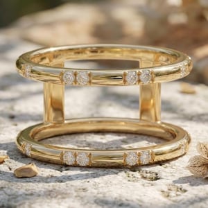 Puede incluir: Un anillo dorado con dos bandas paralelas conectadas por barras verticales. Cada banda está adornada con pequeñas piedras brillantes. El anillo se encuentra sobre una superficie texturizada de color claro, con hojas secas en el fondo.