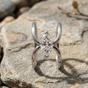 Puede incluir: Un anillo de tono plateado con una piedra preciosa transparente de corte marquesa situada entre dos bandas curvas y paralelas que se extienden hacia arriba y hacia abajo desde la piedra central, creando un diseño abierto y simétrico con acabado pulido.