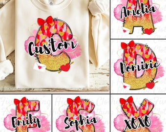 Könnte beinhalten: Ein weißer Pullover mit einem goldenen Glitzerbuchstaben und einer roten Schleife. Der Buchstabe ist von rosa und roten Aquarellherzen umgeben. Der Text "Custom" ist auf dem Pullover geschrieben.