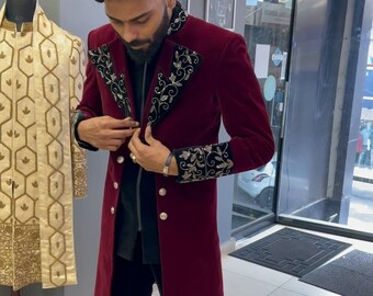 Puede incluir: Un abrigo de terciopelo burdeos con detalles bordados en negro en las solapas y puños. El abrigo presenta un diseño cruzado con botones dorados. La persona lleva pantalones negros y zapatos burdeos. Una prenda de color crema está en un maniquí en el fondo.