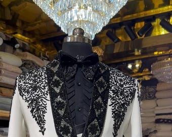 Puede incluir: Chaqueta de esmoquin blanca con adornos de cuentas negras en las solapas, hombros y puños. La chaqueta tiene ribetes y botones negros. Se ve una pajarita negra y una camisa negra.
