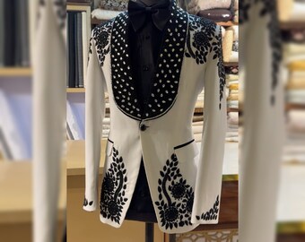 Puede incluir: Una chaqueta blanca con bordados florales negros y un cuello de cuentas. La chaqueta tiene un cierre de botón único y una solapa de satén negro.