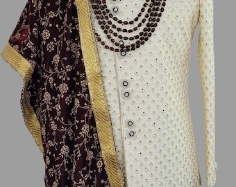 May include: A white embroidered men's sherwani with a maroon floral print shawl. The sherwani has a high collar and a button closure. The shawl has a gold border and is draped over the left shoulder. The man is wearing a maroon turban with a gold and red jewel.
