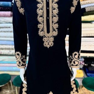 May include: A black tunic with elaborate gold embroidery. The long-sleeved garment showcases detailed floral and scrollwork patterns on the collar, cuffs, and front. It has a mandarin collar and a button-down front. The embroidery is ornate and adds a touch of sophistication.