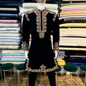May include: A black tunic and trousers ensemble with gold embroidery, presented on a mannequin. The tunic has detailed gold patterns around the collar, front, cuffs, and hem. The mannequin is positioned in front of shelves filled with various coloured fabrics.