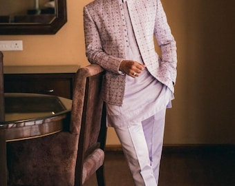 Puede incluir: Un hombre con un blazer estampado en lavanda y pantalones a juego con una camisa blanca de cuello debajo. Está de pie junto a una silla marrón.