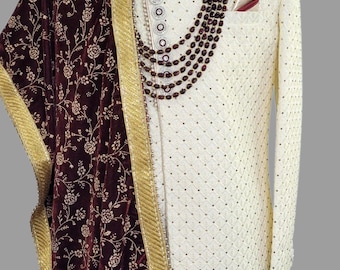 May include: A white patterned kurta with a maroon velvet shawl with gold trim. The shawl has a floral pattern. The kurta has a pearl necklace and a maroon pocket square.