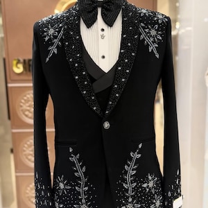 Puede incluir: Un esmoquin negro con intrincados bordados plateados en las solapas, mangas y parte delantera. El traje incluye una pajarita a juego, una camisa blanca y un chaleco negro. La ropa formal se exhibe en un maniquí.