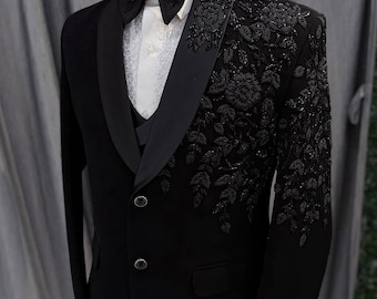May include: A formal black tuxedo with a satin lapel, bow tie, and vest. The left side of the jacket features intricate floral embroidery. The suit is displayed on a mannequin, showcasing the tailored fit and design.