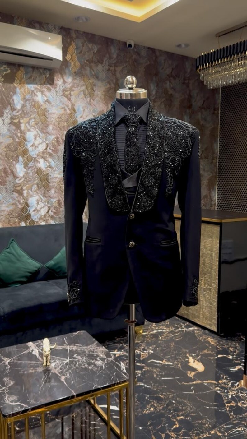 Puede incluir: Un traje negro con intrincados adornos de cuentas en las solapas y pu&ntilde;os. El traje incluye una corbata y chaleco a juego, exhibido en un maniqu&iacute;. Es una prenda formal.