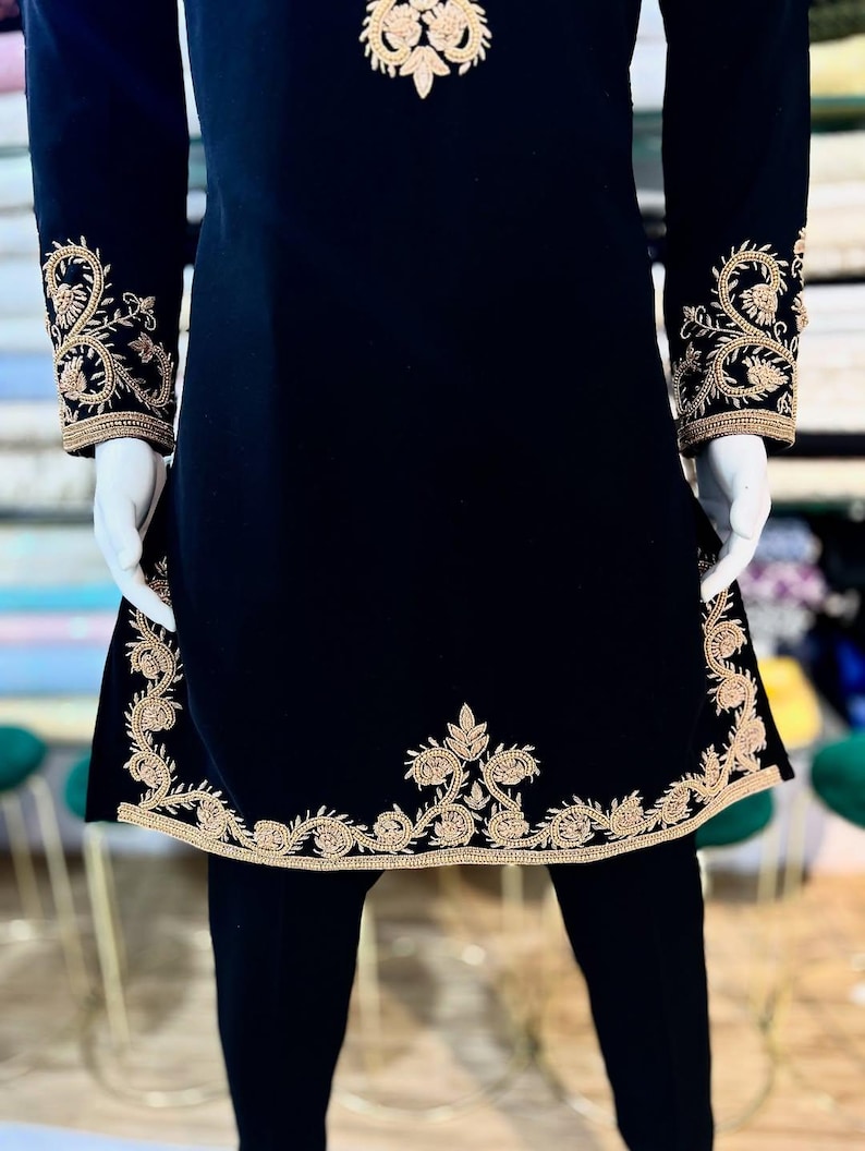 May include: Black tunic with gold embroidery. The tunic has detailed gold floral embroidery on the cuffs, hem, and upper chest. The sleeves are long, and the tunic has a straight, slightly A-line shape. The garment is displayed on a mannequin.