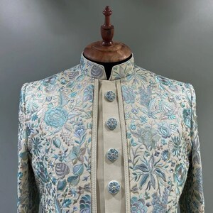 May include: A cream-colored jacket with a blue and silver floral embroidery pattern. The jacket has a mandarin collar and a button closure.