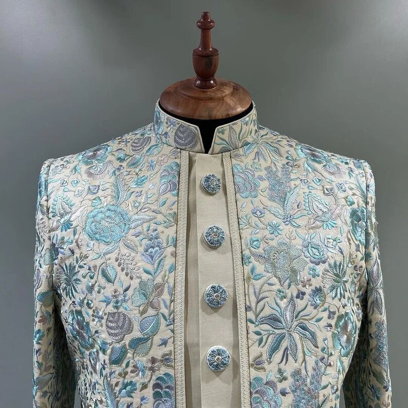 May include: A cream-colored jacket with a blue and silver floral embroidery pattern. The jacket has a mandarin collar and a button closure.