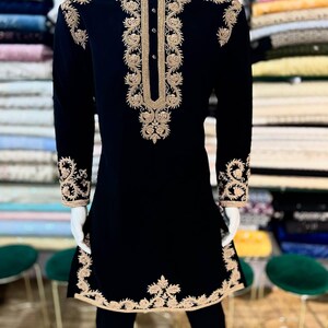 May include: Black embroidered tunic and trousers. The tunic has detailed gold floral embroidery on the collar, front, cuffs, and hem. The long-sleeved tunic features a mandarin collar and a button-down front. The outfit is displayed on a mannequin.