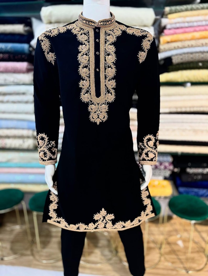 May include: Black embroidered tunic and trousers. The tunic has detailed gold floral embroidery on the collar, front, cuffs, and hem. The long-sleeved tunic features a mandarin collar and a button-down front. The outfit is displayed on a mannequin.