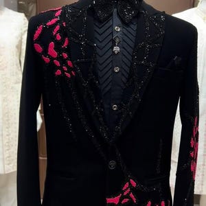 May include: Black tuxedo jacket with a black bow tie and a black dress shirt. The jacket has a velvet texture and is embellished with pink and black sequin designs. The shirt has a pleated front and a decorative brooch.