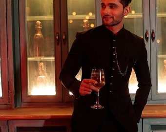 May include: A man wearing a black suit jacket with a chain detail stands in a dimly lit room. He holds a glass of white wine in his right hand and looks to the left of the frame.