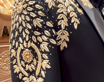 Puede incluir: Chaqueta formal negra con intrincados bordados dorados en el hombro y la manga. El diseño presenta un patrón de hojas y flores. La chaqueta se exhibe en un maniquí, mostrando el ajuste a medida y el diseño elegante.