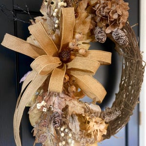 May include: A decorative fall wreath featuring a large burlap bow, dried leaves, and pinecones. The wreath is made of woven twigs and adorned with various autumnal elements in shades of brown, beige, and gold. Perfect for seasonal home decor.