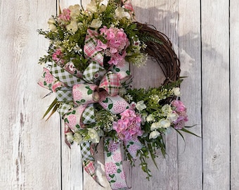 May include: A decorative wreath featuring a grapevine base adorned with pink hydrangeas, white flowers, and greenery. A large bow with pink floral and green gingham patterns adds a charming touch. The wreath is displayed against a white wooden background.