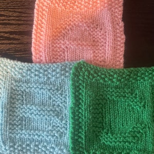 Könnte beinhalten: Drei gestrickte Quadrate in verschiedenen Farben: rosa, hellblau und grün. Jedes Quadrat hat ein anderes Strickmuster.