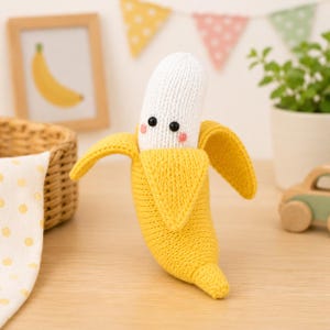 Peut inclure: Un jouet banane tricoté présentant une partie supérieure blanche pelée avec deux yeux en perles noires et de petites joues roses brodées, enveloppé dans une peau jaune vif. La banane a une texture douce et des mailles tricotées visibles.