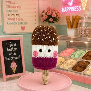 May include: A knitted ice cream bar toy with a brown top featuring white embroidered sprinkles, a white middle section with black bead eyes, pink cheeks, and a dark purple bottom, all attached to a tan stick and resting on a pink pedestal stand.