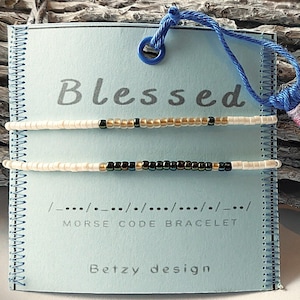 May include: Two beaded bracelets on a blue card with the word "Blessed" and the text "Morse Code Bracelet" and "Betzy design". The card has a blue tassel attached.