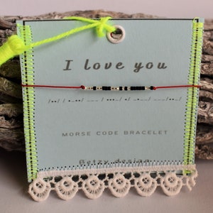 Könnte beinhalten: Ein Morsecode-Armband mit schwarzen und silbernen Perlen auf einer roten Schnur, präsentiert auf einer hellblauen Karte. Auf der Karte steht "I love you" und "Morse Code Bracelet". Die Karte ist mit neongrünen Nähten und weißer Spitze verziert.