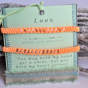 Könnte beinhalten: Zwei neonorangefarbene Morsecode-Armbänder werden auf einer hellgrünen Karte mit dem Wort "Love" oben präsentiert. Die Karte enthält auch den Text "Morse Code Bracelet" und ein Zitat. Eine hellgrüne Quaste hängt daran.
