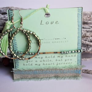 Op de afbeelding: Hoepelvormige oorbellen met groene kralen en een bijpassende kwast, gepresenteerd met een Morsecode armband en een kaart met het woord "Love". De kaart bevat de quote: "Je mag mijn hand even vasthouden, maar je houdt mijn hart voor altijd."