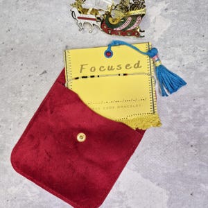Könnte beinhalten: Ein rotes Samtetui mit einem goldenen Druckknopf enthält eine gelbe Karte mit dem Wort "Focused" und ein Morsecode-Armband. Ein blauer Quastenanhänger und ein dekorativer Weihnachtsschlitten sind ebenfalls zu sehen.