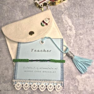 May include: A green and black Morse code bracelet on a light blue card that says "Teacher". The card is attached to a light blue tassel and is inside a beige pouch. The bracelet is made of woven green thread and black beads.