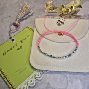 Pode incluir: Uma pulseira rosa trançada com contas prateadas e azuis, em uma bolsa branca. Uma etiqueta verde com as palavras "Never give up" e uma borla também estão presentes. Um enfeite de trenó dourado está no fundo.
