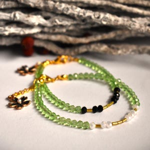 May include: Two delicate bracelets with green and black beads, accented with gold and white flower charms.