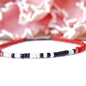 May include: Red beaded bracelet with a central pattern of black and white beads. The bracelet is strung on a red cord and has gold-coloured accents. The background features a blurred pink floral arrangement.