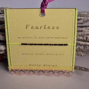 Peut inclure: Bracelet en code Morse sur une carte jaune avec le mot "Fearless" imprimé. Le bracelet est noir et monté sur un cordon jaune fluo. La carte a une bordure en dentelle rose et un cordon violet pour l'accrocher.