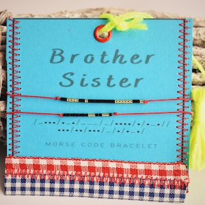 Może przedstawiać: Niebieska karta z napisami "Brother Sister" i dwiema bransoletkami z kodem Morse'a. Bransoletki mają czarne i złote koraliki na czerwonych sznurkach. Karta ma czerwoną i białą wstążkę w kratkę na dole oraz napis "MORSE CODE BRACELET".