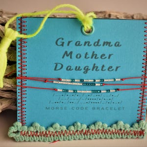 May include: A turquoise blue card with red stitching around the edges. The card has the words "Grandma Mother Daughter" printed in white.  Three red strings with silver and turquoise beads are attached to the card. The text "Morse Code Bracelet" is printed below the beads.