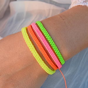 Könnte beinhalten: Vier farbenfrohe, gewebte Armbänder in Neon-Gelb, Orange, Pink und Grün sind auf einem Handgelenk gestapelt. Die Armbänder bestehen aus einem strukturierten Material. Ein lebendiges Accessoire für jedes Outfit.