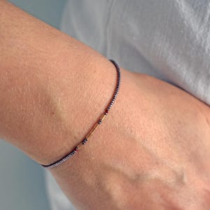 May include: A delicate bracelet with a dark beaded chain. The bracelet features gold-colored and red accents. The bracelet is worn on a wrist, with a white shirt in the background.